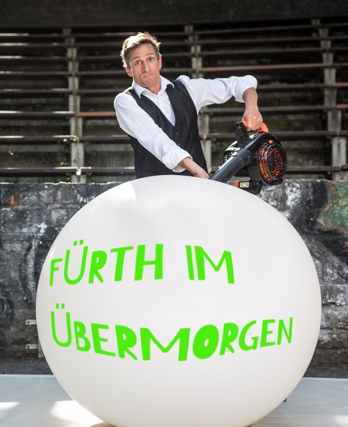 Comedian Markus Just pumpt einen Riesenballon auf, auf dem Fürth im Übermorgen steht.