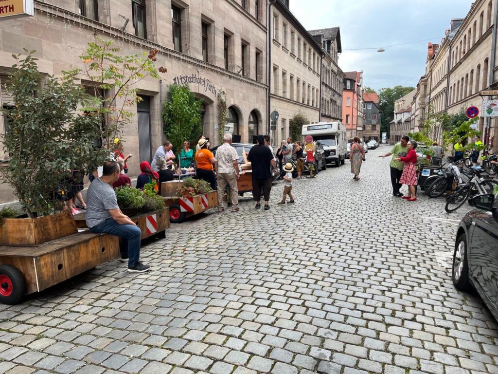 Menschen sitzen auf den Bänken von Pflanztrgen, Unterhalten sich, genießen den Sommer in einer gepflasterten Altstadtstraße.