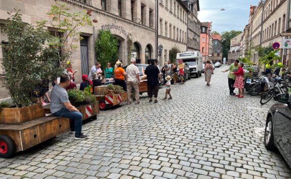 Menschen sitzen auf den Bänken von Pflanztrgen, Unterhalten sich, genießen den Sommer in einer gepflasterten Altstadtstraße.