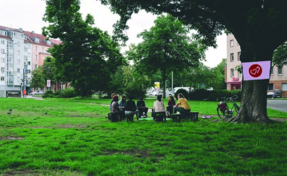 Eine Gruppe Menschen sitzt unter einem Stadtbaum zusammen und unterhält sich.